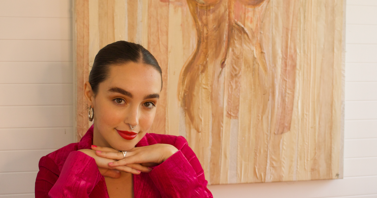 photo fo woman with hands under her chin, she has a bright pink jacket on and is sitting in front of a painting.